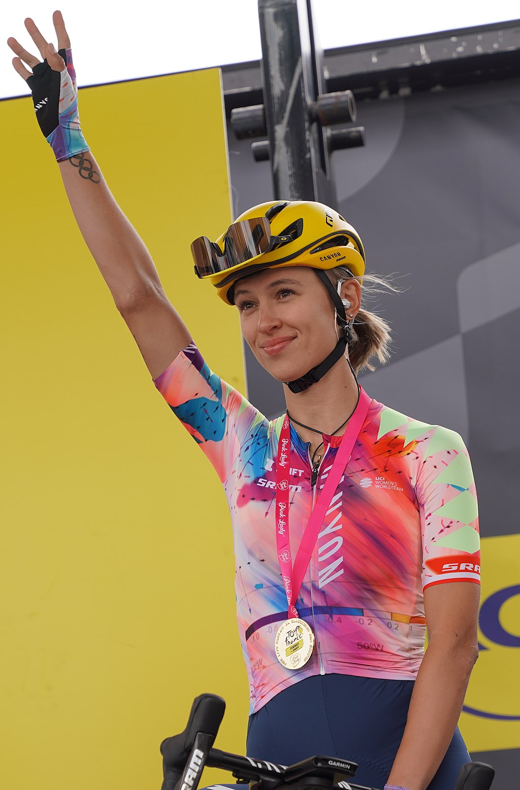 Katarzyna Niewiadoma ubrana w kolorową koszulkę i żółty kask podnosi lewą rękę, trzymając medal na szyi, stoi obok żółto-szarego tła.