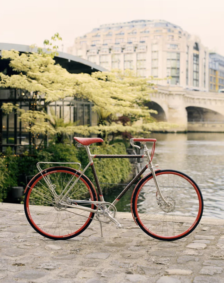 Zabytkowy rower Maison Tamboite Paris zaparkowany na brukowanym brzegu rzeki z mostem i budynkami w tle.
