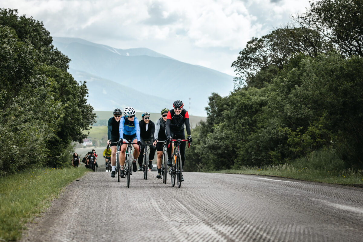 Grupa rowerzystów w odzieży sportowej jadących wiejską drogą z górami w tle w pochmurny dzień.