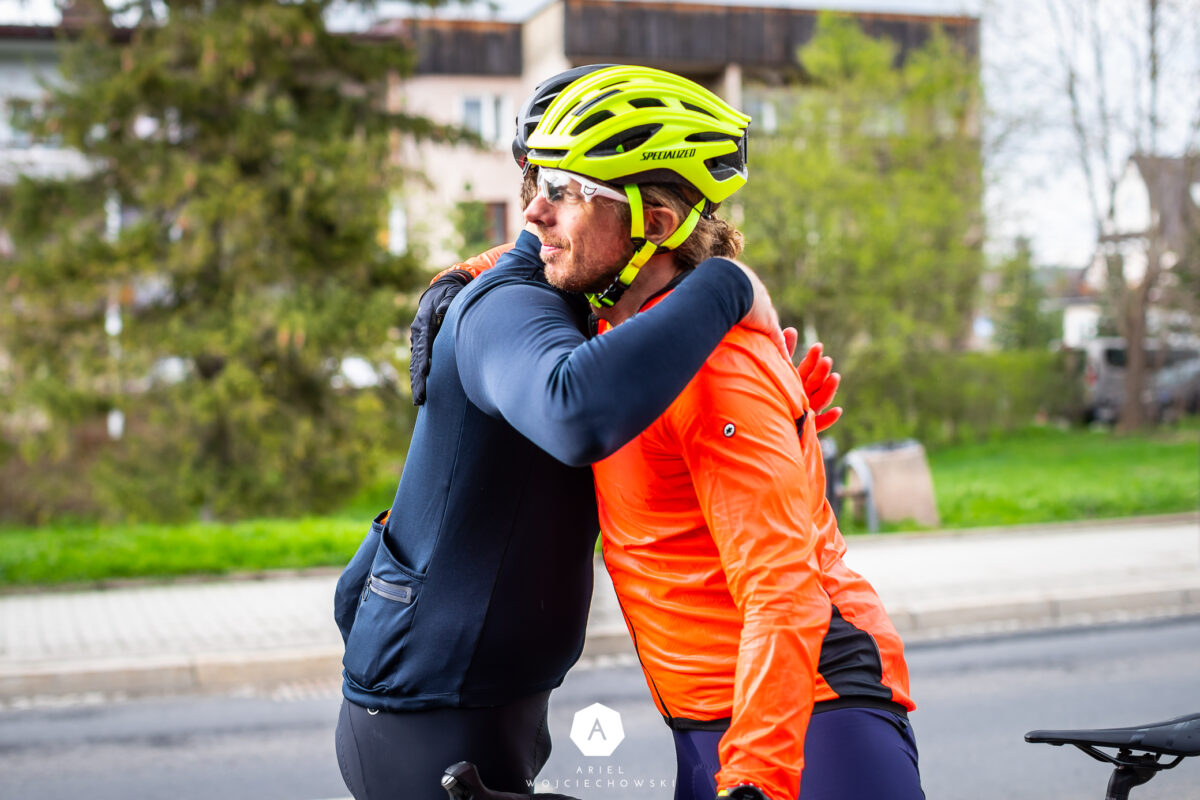 Dwóch rowerzystów przytulających się na ulicy miejskiej, jeden w żółtym kasku, drugi w pomarańczowej kurtce.