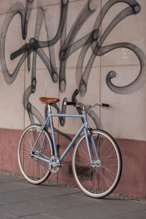 Niebieski rower z brązowym siodełkiem stoi zaparkowany przy beżowej ścianie pokrytej czarnym graffiti.