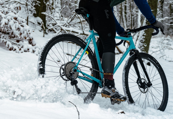 Rowerzysta jedzie na turkusowym rowerze po pokrytej śniegiem leśnej ścieżce.