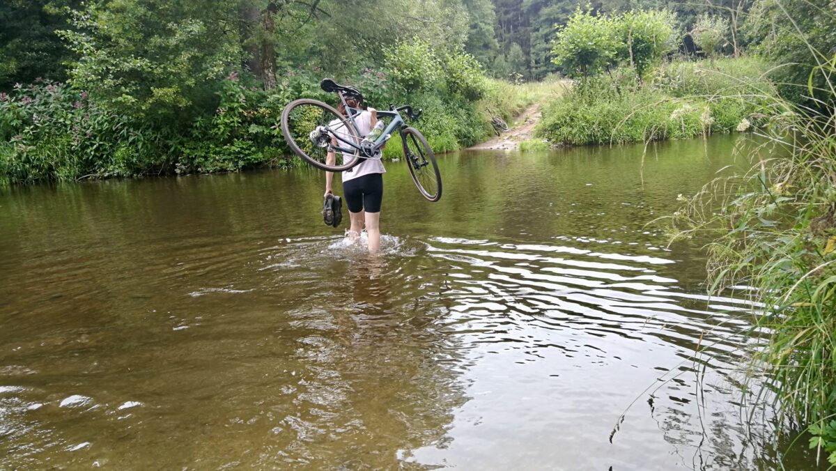 Podczas wyścigów gravelowych czasem trasa zamienia się w rzekę. Tutaj zawodniczka przenosi rower i buty.