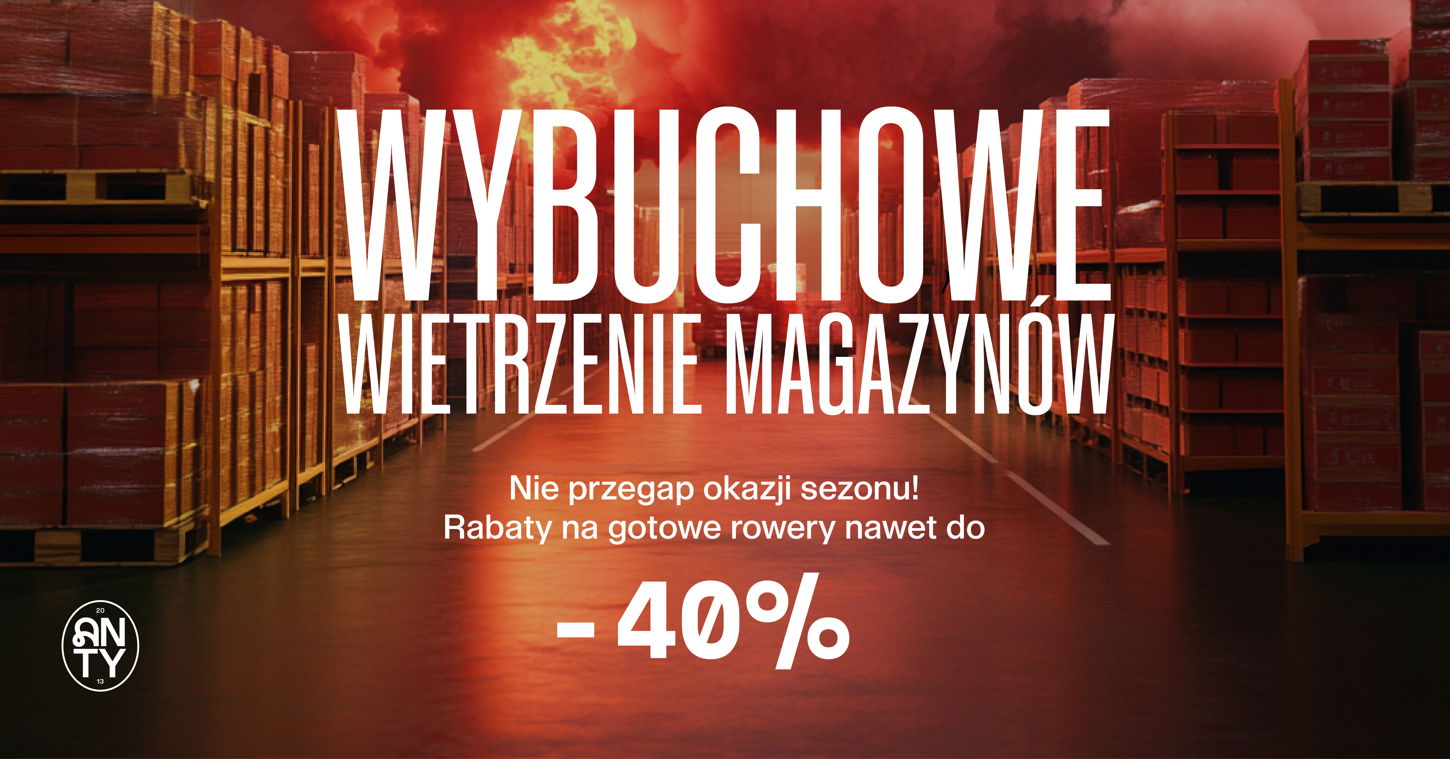 Dramatyczna scena magazynowa z ułożonymi półkami i ognistym niebem. Tekst pogrubiony brzmi: „WyBUCHOWE WIETRZENIE MAGAZYNÓW” i „Rabaty na gotowe rowery nawet do -40%”.