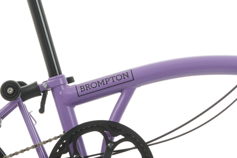 Zbliżenie na fioletową ramę roweru Brompton przedstawiające część łańcucha, pedału i tylnego koła.