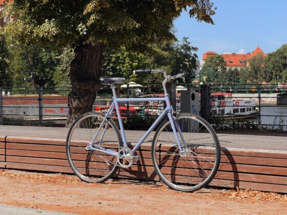 Antymateria Miasto ON - błękitny 56cm zaparkowana jest na drewnianej ławce przy rzece w Mieście On. W słoneczny dzień w tle widać drzewa i most.