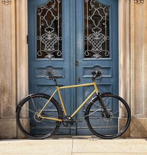 Żółty rower z Antymaterią Pasek - rower paskowy roz. L Kameleon zielono-niebieski stoi przed ozdobnymi niebieskimi podwójnymi drzwiami ozdobionymi ozdobnymi okuciami na kamiennym budynku.