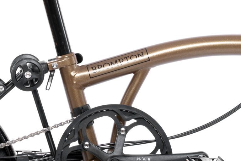 Zbliżenie na brązową ramę Brompton P-line Urban M4R Bronze Sky, skupiając się na środkowej części z logo Brompton, części łańcucha, pedałów i mechanizmu zmiany biegów. Misternie zaprojektowane przekładnie i eleganckie wykończenie ukazują zaangażowanie firmy Brompton w precyzyjną inżynierię i stylową funkcjonalność.