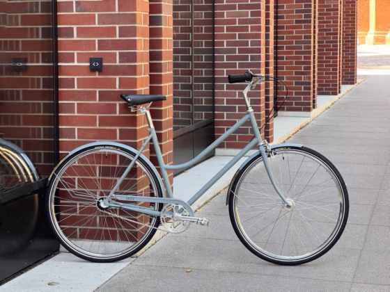 Na chodniku obok ceglanego budynku zaparkowany jest srebrny rower Antymateria Miasto ONA -Szary mat XL o minimalistycznym designie.