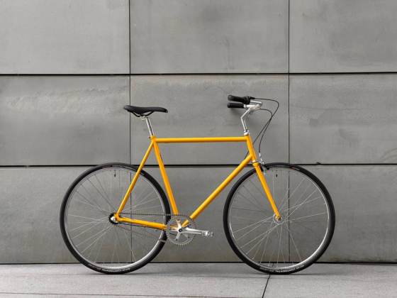 Żółty rower Antymateria Miasto ON - zółty 59cm z czarnym siodełkiem i kierownicą oparty jest o szarą betonową ścianę. Rower ma minimalistyczny design i cienkie opony.