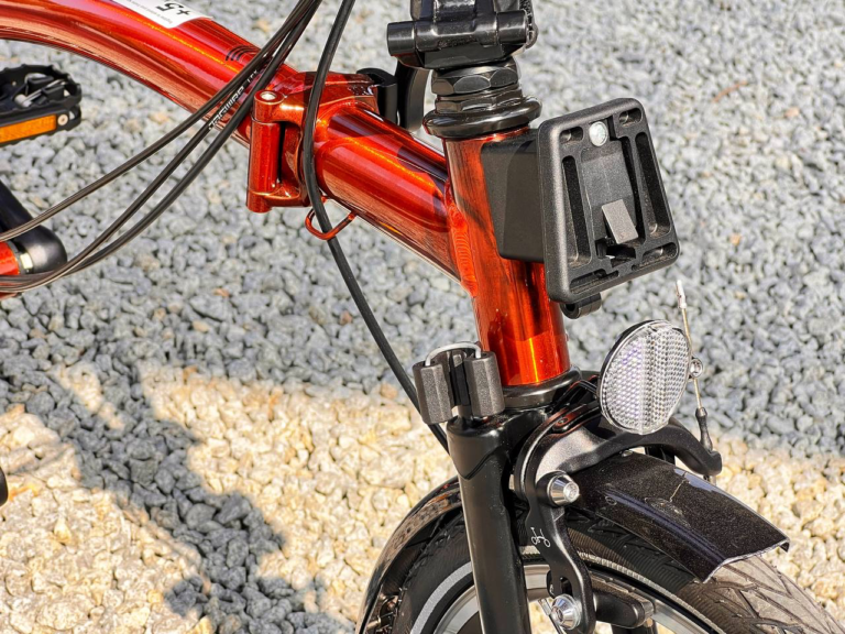 Zbliżenie przedniej ramy i koła składanego roweru Brompton C-line M6R Flame Lacquer - Black Edition, ukazujące reflektor, elementy hamulca i uchwyt montażowy na tle żwiru.