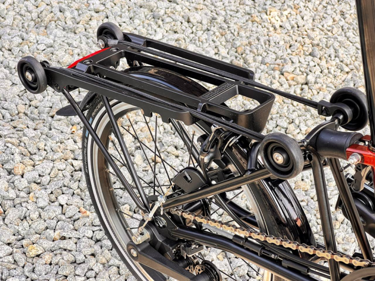 Zbliżenie złożonego roweru Brompton C-line M6R Flame Lacquer - Black z małymi kołami na żwirowej nawierzchni, ukazujące jego kompaktową konstrukcję i mechanizm składania.