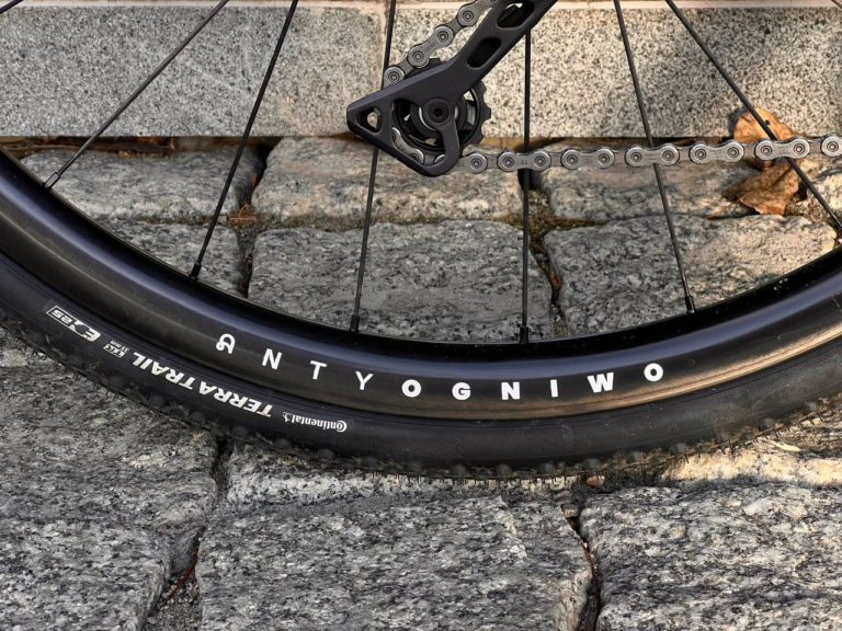 Opona rowerowa na brukowanej nawierzchni z napisem „ANTYOGNIWO” i „TERRA TRAIL” z widocznym segmentem łańcucha i tylną przerzutką, uosabiająca model Anty Gravel CRE M - GRX400/600 - Czarny mat — idealny na każdą przygodę na szutrze CRE.