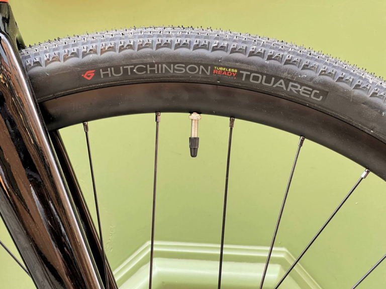 Zbliżenie koła rowerowego z oponą Hutchinson Touareg i widocznym oznaczeniem „tubeless ready”. Ten komponent Antymateria Gravel - pomarańczowy metalic M (54 cm) jest osadzony na solidnej zielonej ścianie, co podkreśla jego solidną konstrukcję.