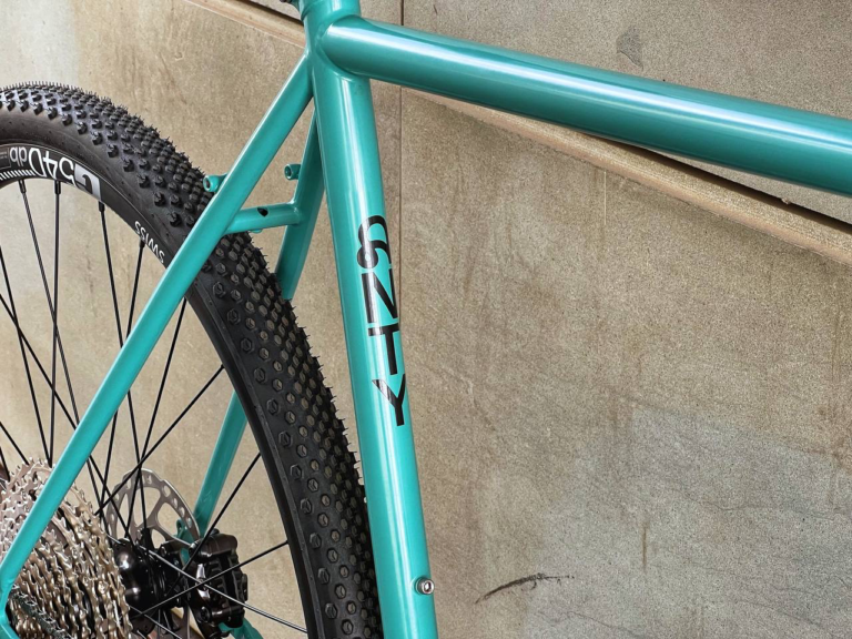 Zbliżenie na niebieską ramę roweru Antymateria Gravel Rock Shox w rozmiarze L (57 cm) z widoczną na rurze podsiodłowej nazwą marki „SNTY”. Na zdjęciu widać również część tylnej opony i kasetę przerzutek.