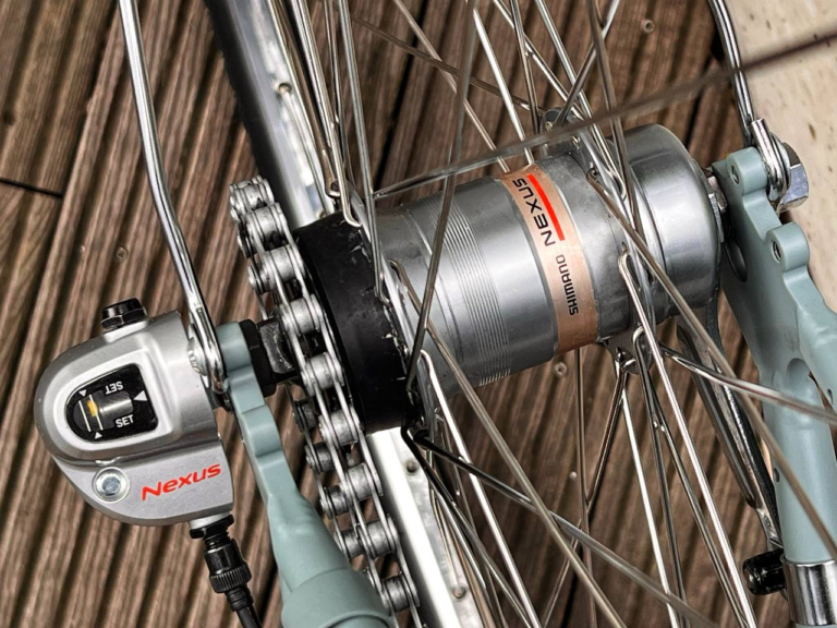 Zbliżenie piasty rowerowej Shimano Nexus z widocznymi szprychami i linką przerzutki w rowerze Antymateria Miasto ONA z koszem - granatowa S (Kopia). W tle znajduje się drewniany pokład.