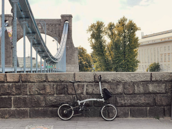 Rower Brompton P-line M4L Lunar Ice zaparkowany przy kamiennym murze z łukowatym mostem i drzewami w tle, w pochmurny dzień.