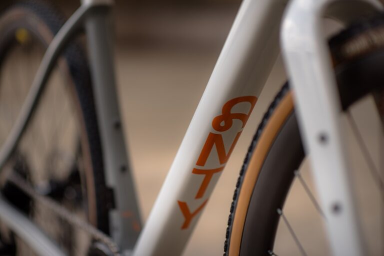 Zbliżenie na ramę roweru z odważnym pomarańczowym napisem „&ty”, skupiające się na teksturowanych szczegółach opony i logo.