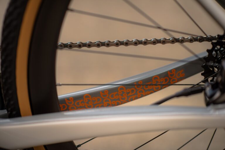 Zbliżenie łańcucha rowerowego i kół zębatych, przedstawiające szczegółowy pomarańczowy wzór na białej ramce.