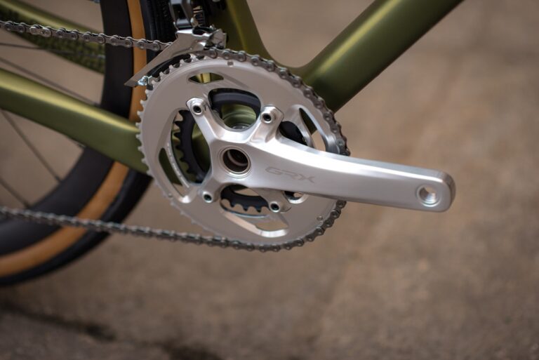 Zbliżenie łańcucha i tarczy nowoczesnego roweru, oznaczone „grx”, przymocowane do oliwkowozielonej ramy z częściowym widokiem na brązową oponę.