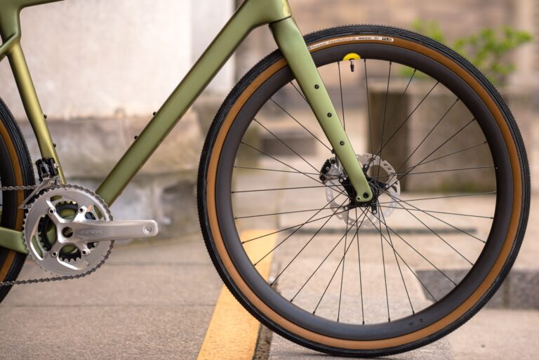 Zbliżenie zielonego roweru, skupiającego się na przednim kole, pedale i części ramy, zaparkowanego na betonowej powierzchni.
