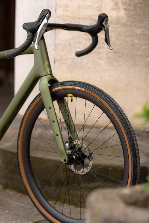 Zbliżenie na zielony rower szosowy oparty o beżową ścianę, skupiający się na kierownicy, przednim kole i oponie.