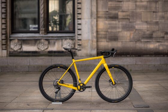 Jasnożółty rower Anty Gravel CRE M (54 cm) - GRX 800 zaparkowany na szarym chodniku pod kamiennym budynkiem z dużymi oknami.