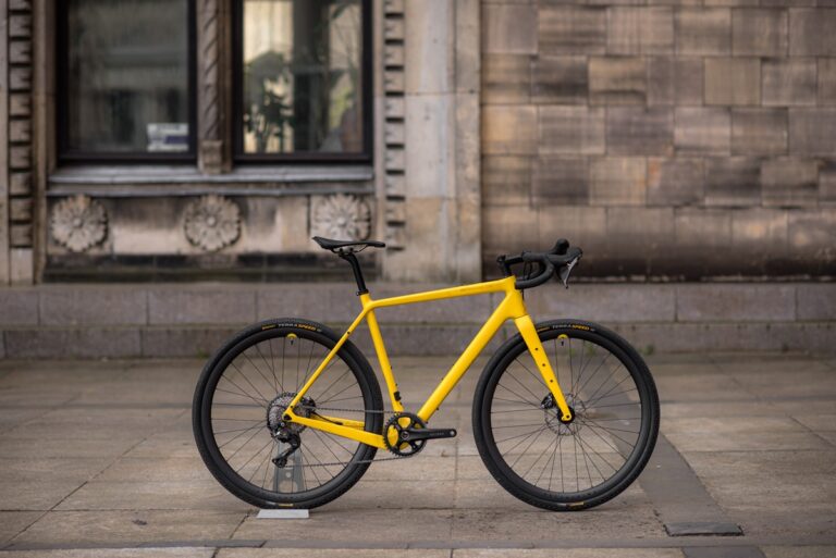 Jasnożółty rower Anty Gravel CRE M (54 cm) - GRX 800 zaparkowany na szarym chodniku pod kamiennym budynkiem z dużymi oknami.