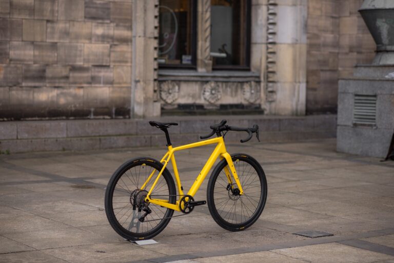 Żółty rower Anty Gravel CRE M (54 cm) - GRX 800 zaparkowany na szarym chodniku pod kamiennym budynkiem z dużymi oknami.