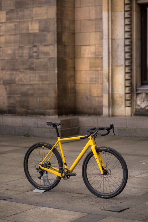 Żółty rower Anty Gravel CRE M (54 cm) zaparkowany pod kamiennym murem przy filarze, na ulicy miejskiej.