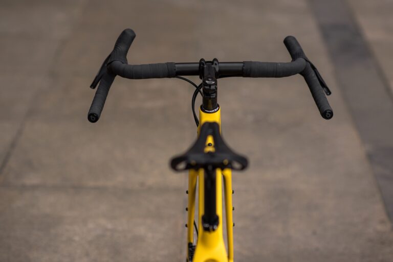 Widok z przodu roweru Anty Gravel CRE M (54 cm) - GRX 800, kierownica i górna rura, skupione, z rozmytym tłem chodnika.
