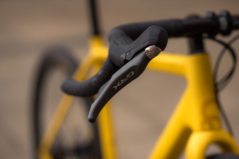 Zbliżenie na czarne hamulce na kierownicy żółtego roweru Anty Gravel CRE M (54 cm) - GRX 800, skupione na dźwigni hamulca z rozmytym tłem.
