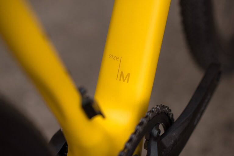 Zbliżenie na żółtą ramę roweru szutrowego Anty Gravel CRE M (54 cm) z metką z rozmiarem „m”.