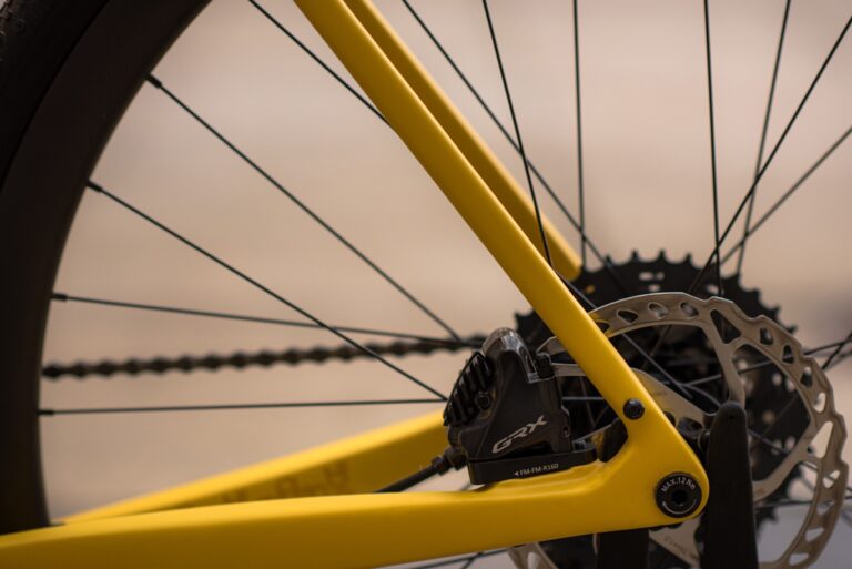 Zbliżenie na żółte tylne koło roweru Anty Gravel CRE M (54 cm) GRX 800, skupione na piaście i szprychach z rozmytym tłem zachodzącego słońca.