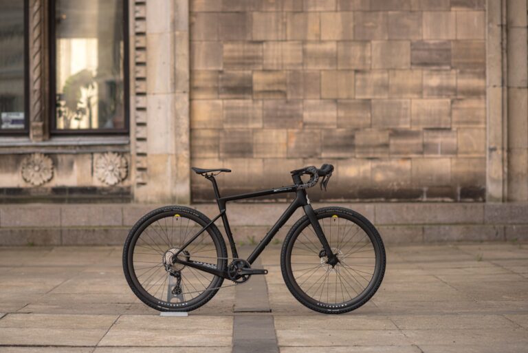 Czarny rower szosowy zaparkowany przed kamiennym budynkiem z dużymi oknami.