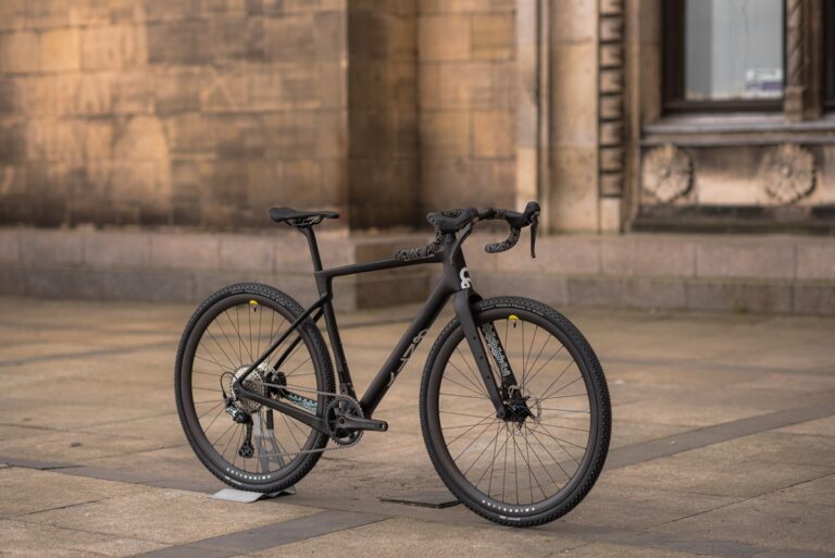 Czarny rower szosowy zaparkowany pod budynkiem z brązowego kamienia, wyposażony w hamulce tarczowe i minimalistyczny design.