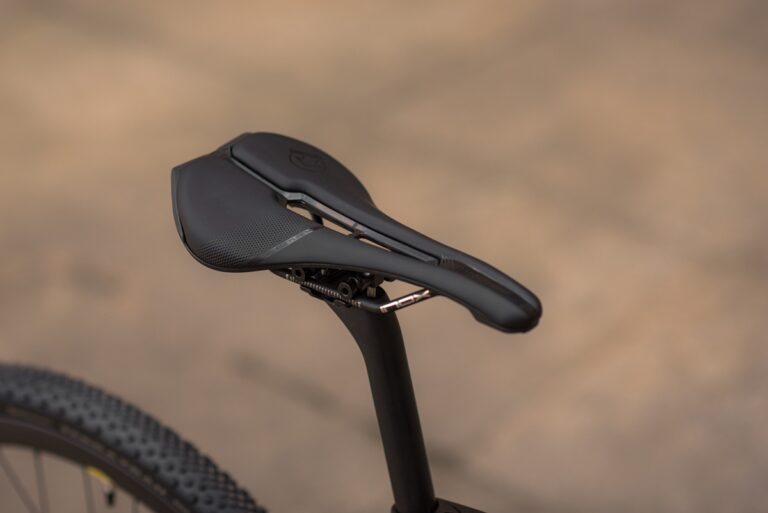 Zbliżenie na nowoczesne siodełko rowerowe o teksturowanej powierzchni i logo marki, zamontowane na sztycy podsiodłowej, z rozmytym tłem.