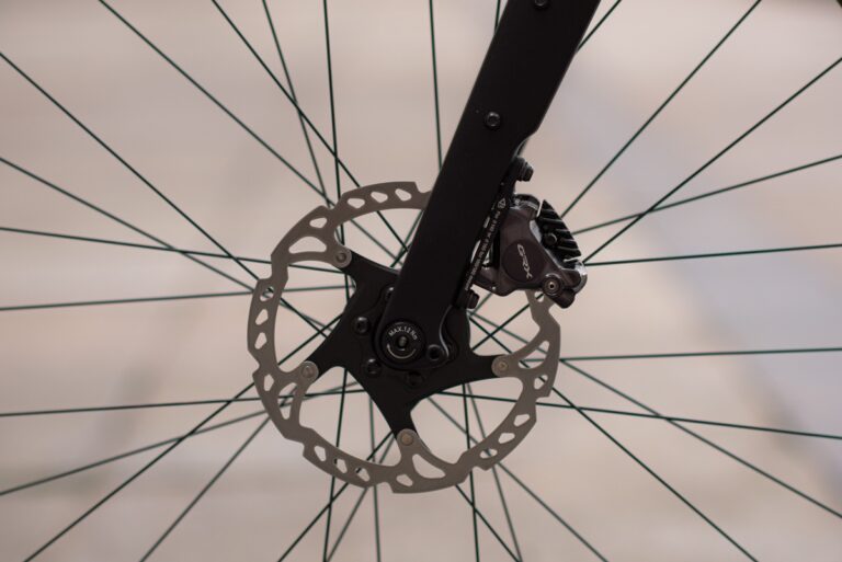 Zbliżenie na hamulec tarczowy roweru i część wzoru szprych przedniego koła, skupiając się głównie na mechanizmie hamulca i tarczy.
