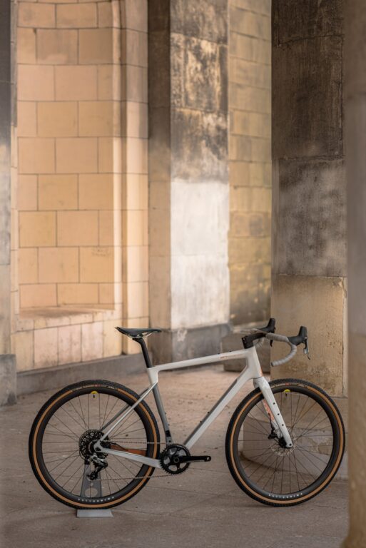 Elegancki biały rower szosowy oparty o kamienną kolumnę w zadaszonym pasażu, oświetlony miękkim, naturalnym światłem.