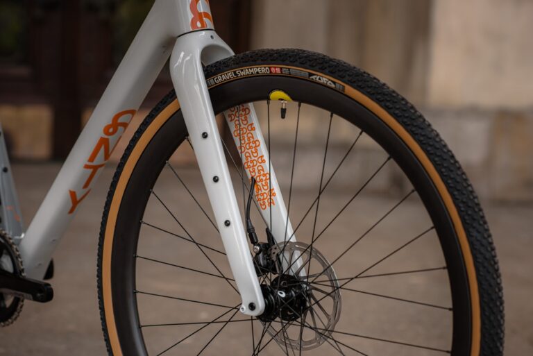 Zbliżenie na przednie koło roweru ze szczegółowym widokiem widelca i opony z napisem „strada bianca” na tle miejskim.