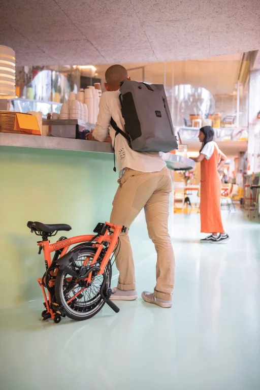 Osoba stoi przy ladzie z plecakiem Torba Brompton Borough Waterproof L - Navy (Kopia) przewieszonym przez ramię, obok złożonego pomarańczowego roweru.