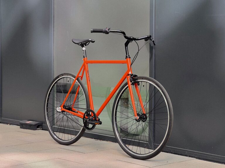 Pomarańczowy rower zaparkowany pod szarą ścianą na chodniku.
