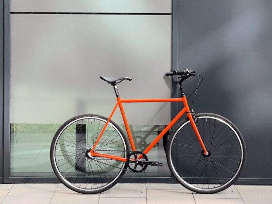 Pomarańczowy rower zaparkowany pod szarą ścianą z dużym odblaskowym oknem po prawej stronie.