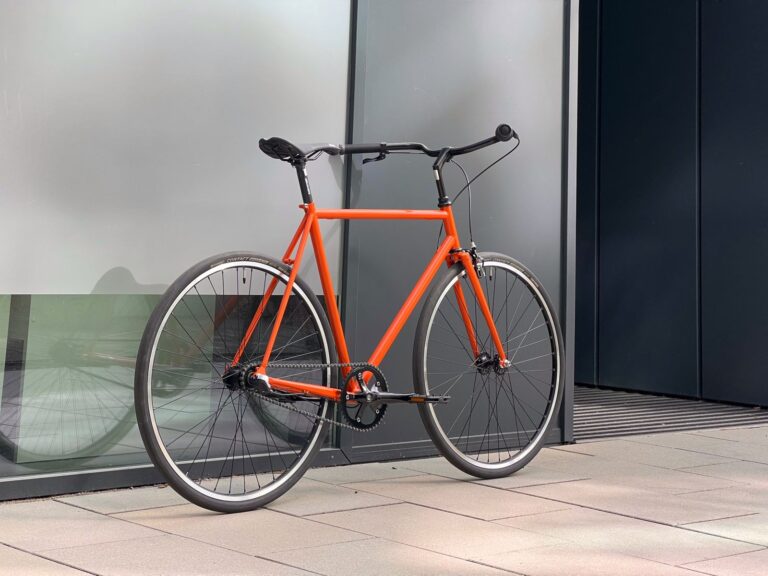 Pomarańczowy rower zaparkowany na tle szarego budynku z dużym oknem, elegancką ramą i czarną kierownicą.