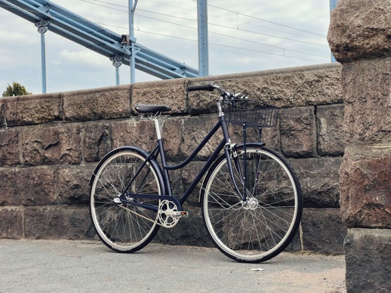 Niebieski rower z przednim koszykiem zaparkowany pod kamienną ścianą, na tle mostu.