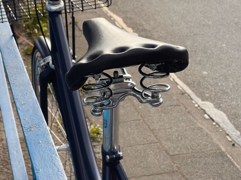 Zbliżenie siodełka rowerowego zamontowanego na metalowym sztycy, z częściowo widoczną niebieską ramą. w tle widać rozmytą ulicę miejską.