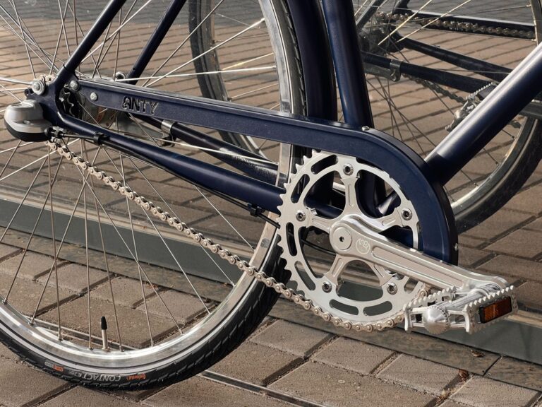 Zbliżenie na tylne koło i pedał niebieskiego roweru, podkreślające mechanizm zmiany biegów i łańcuch, zaparkowane na ceglanej powierzchni.