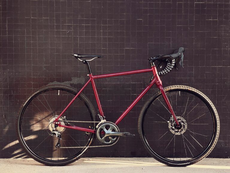 Zdanie z nazwą produktu: Czerwony rower Antymateria All Terrain - na dystanse 53cm opiera się o ciemną ceglaną ścianę, ma obniżoną kierownicę i hamulce tarczowe.
